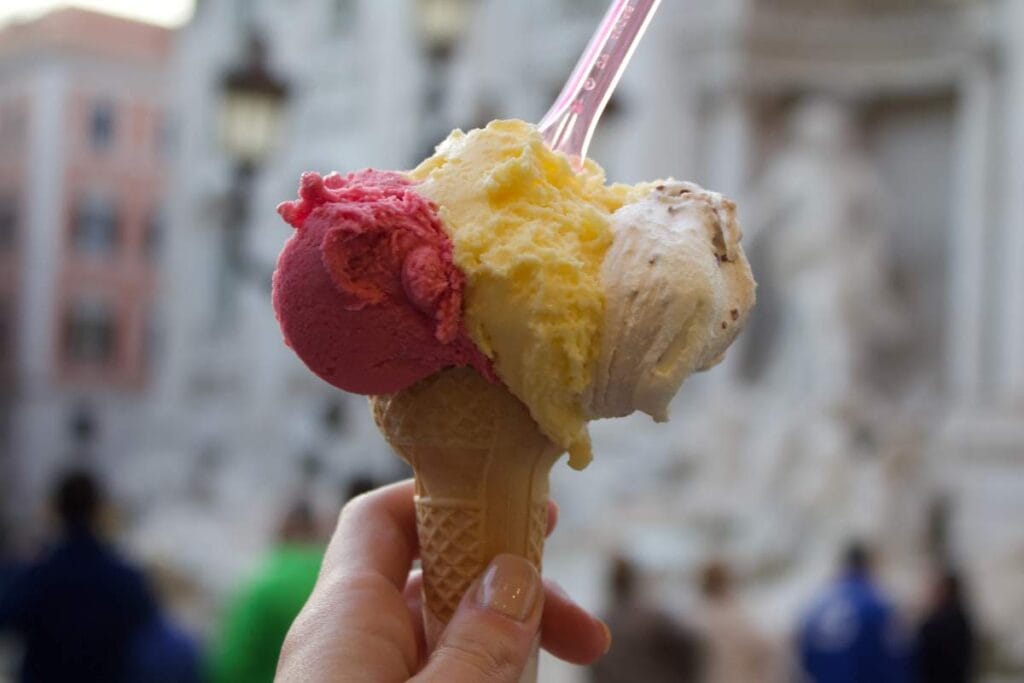 Close-up of a three-scoop gelato cone with a blurred Roman fountain background